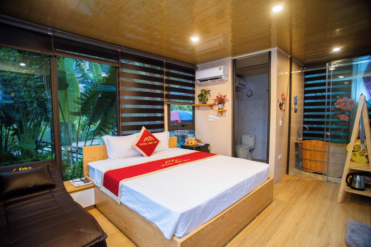 Une chambre d’hôtel moderne dotée d’un grand lit, d’un bureau en bois et offrant une vue sur une végétation luxuriante grâce à de grandes fenêtres du sol au plafond.