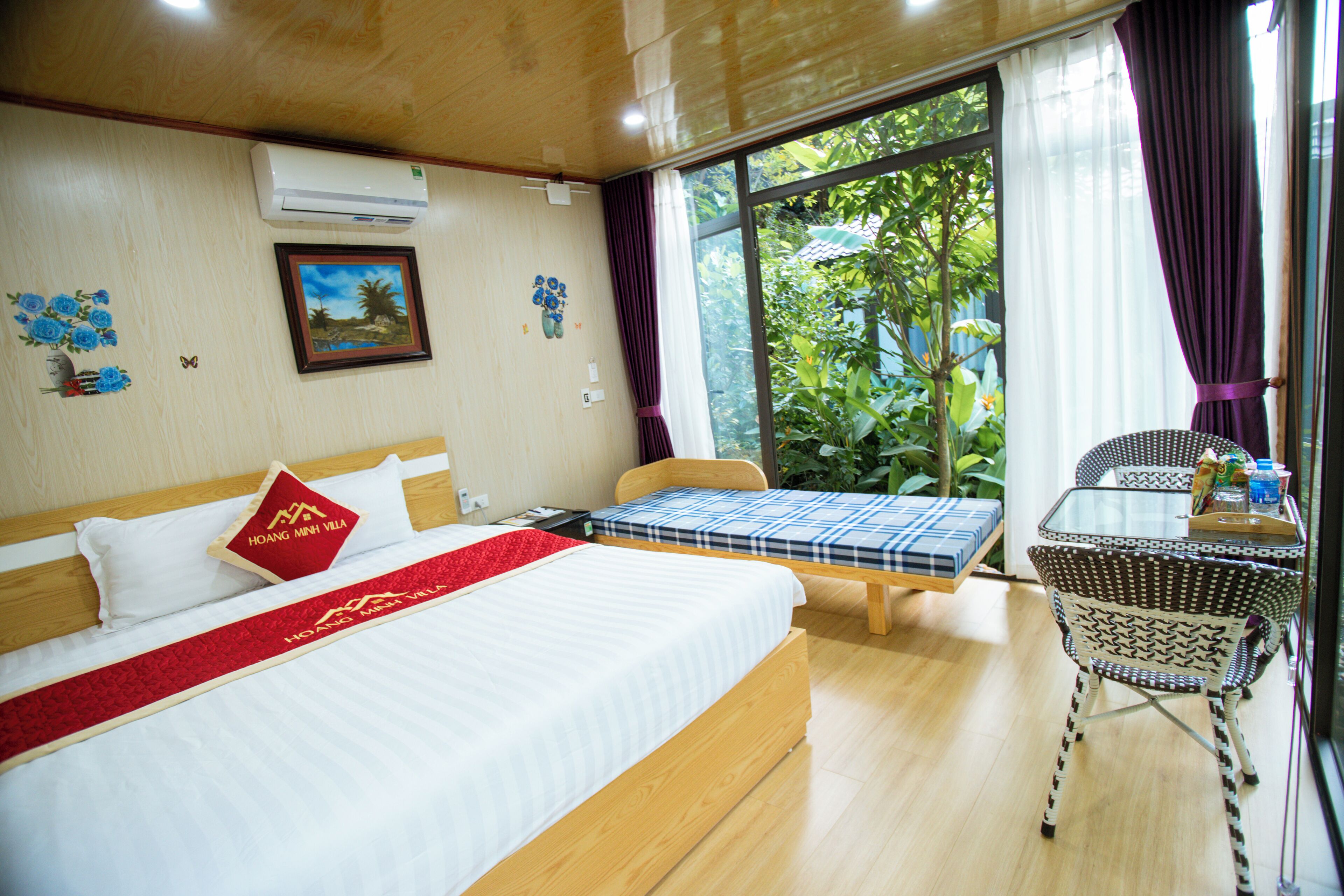 A hotel room with a large bed, a smaller bed, a table, and a chair. There is a window with curtains and a view of greenery outside.