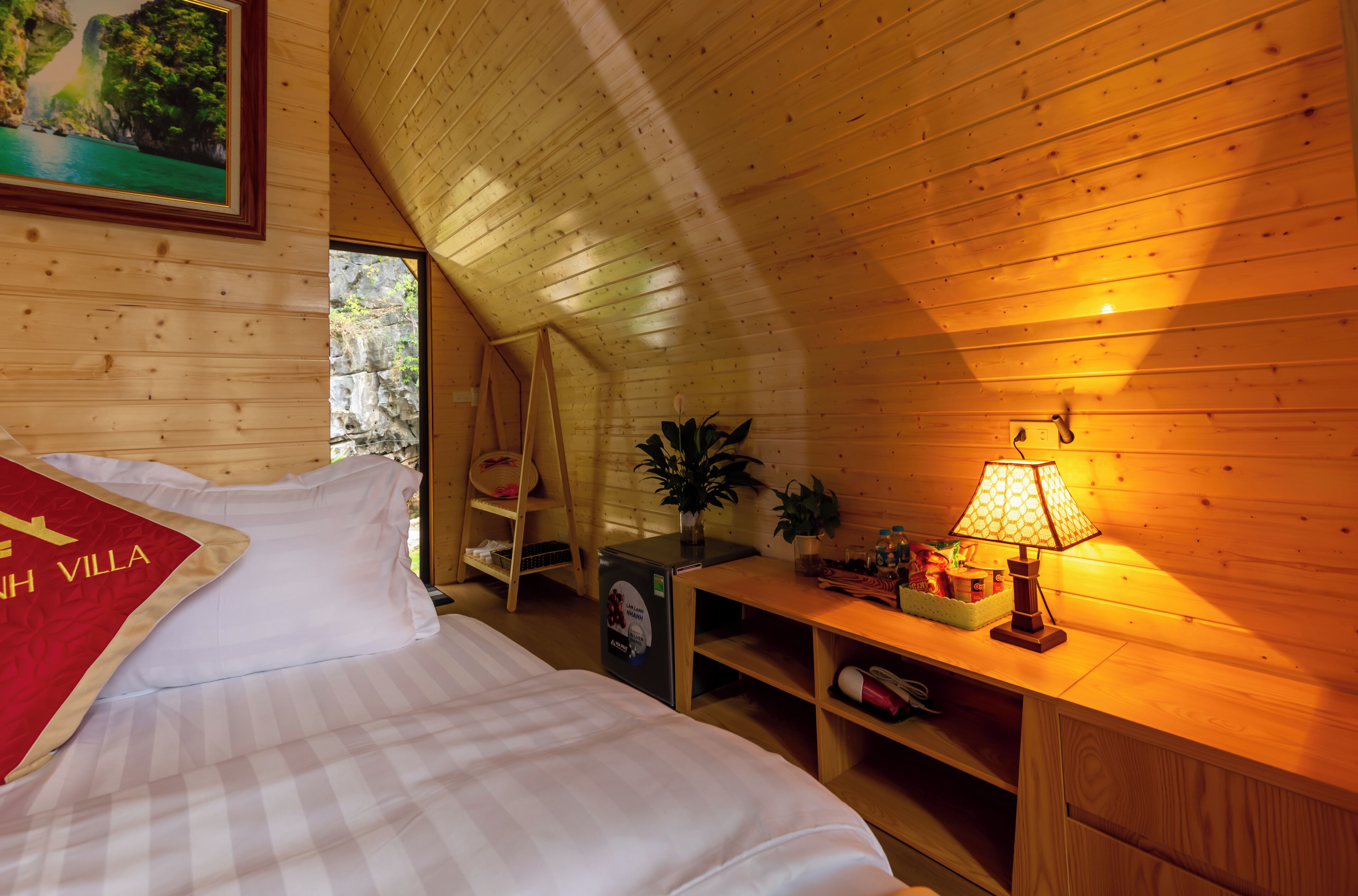 A cozy room with a bed, a desk with a lamp, a shelf with items, and a painting on the wall.