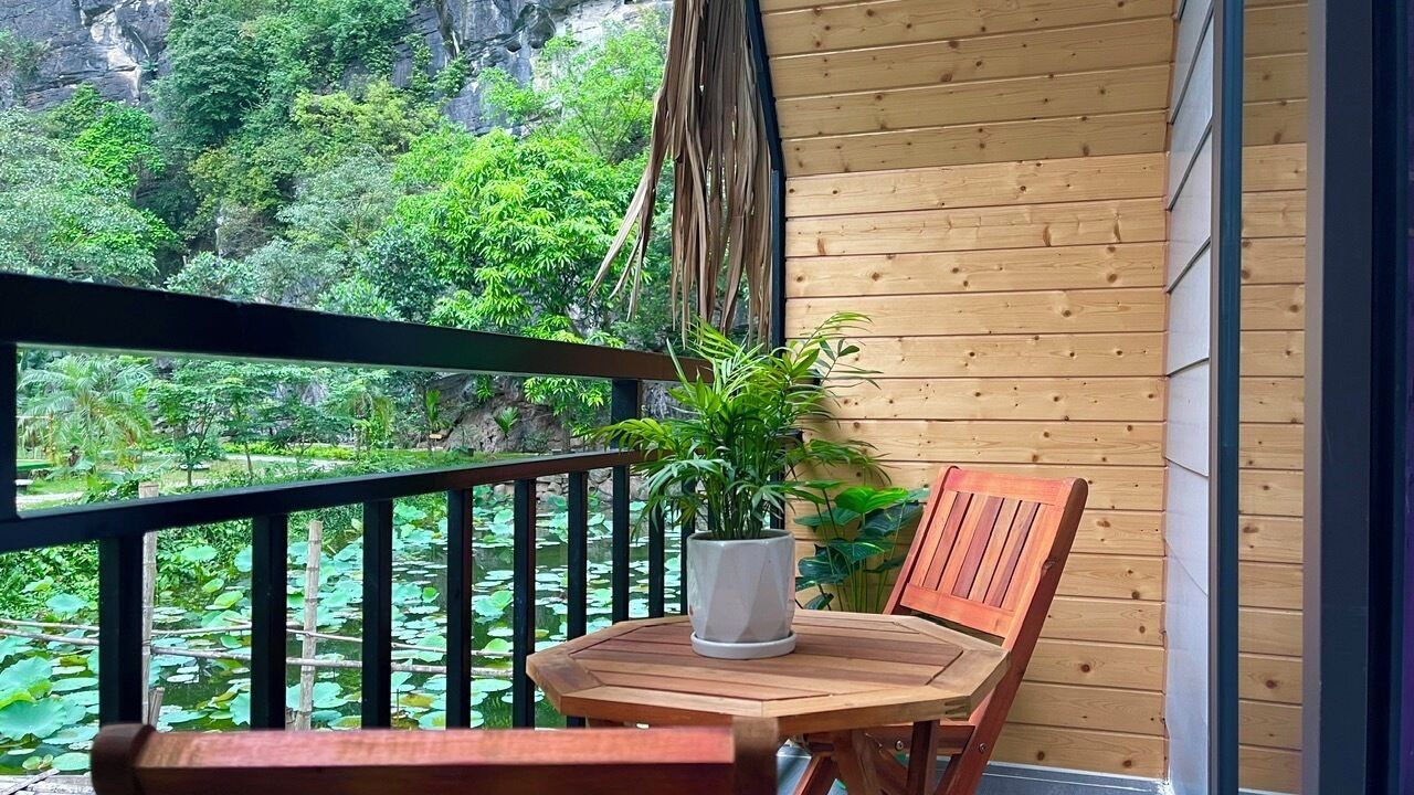 A wooden balcony with a table and chairs, a potted plant, and a view of a lush garden with trees and a rock formation.