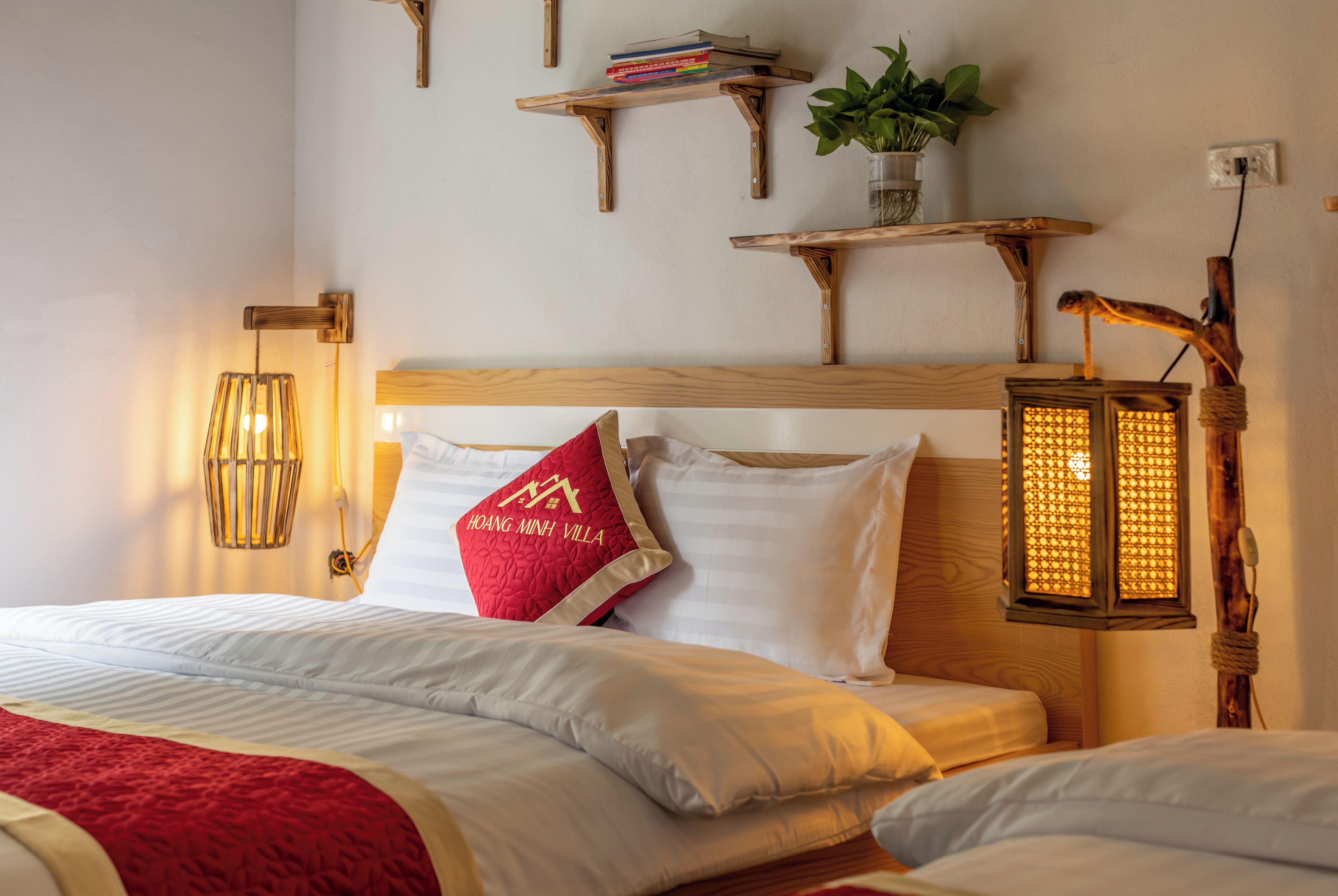 A neatly made bed with white linens, a red pillow, and a wooden headboard. There are two wall-mounted shelves above the bed, one holding books and a potted plant, and the other with a decorative lantern.