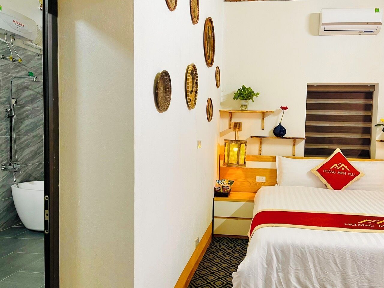 Habitación de hotel con cama, mesita de noche, lámpara, aire acondicionado instalado en la pared y baño con ducha e inodoro.