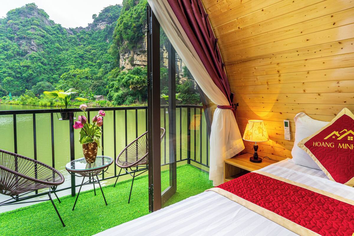 Une chambre d’hôtel comprenant un lit, une table de chevet, une lampe, un oreiller rouge, une fenêtre donnant sur un paysage verdoyant et un lac, ainsi qu’un balcon avec des chaises et une table.