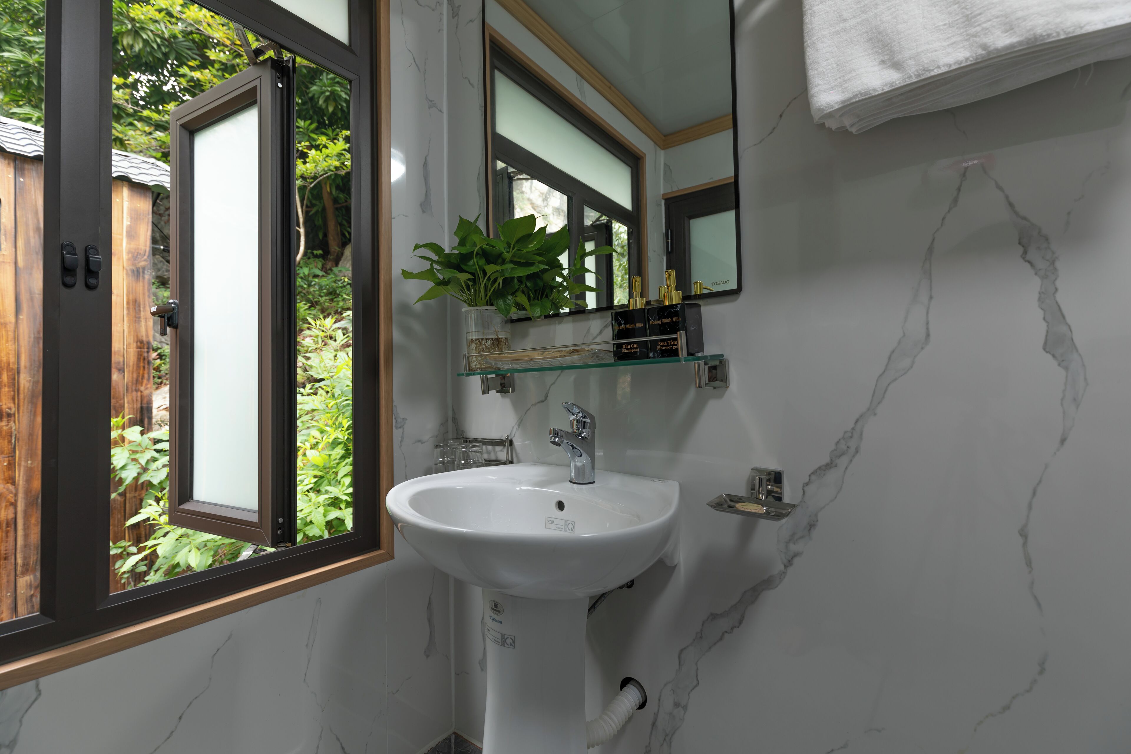 A bathroom with a white sink, a mirror, and a window with a view of greenery outside.