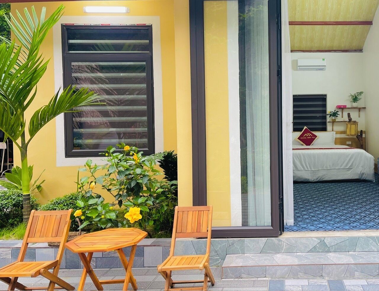 A hotel room with a bed, a small table, and chairs outside. There is a window and a plant near the entrance.