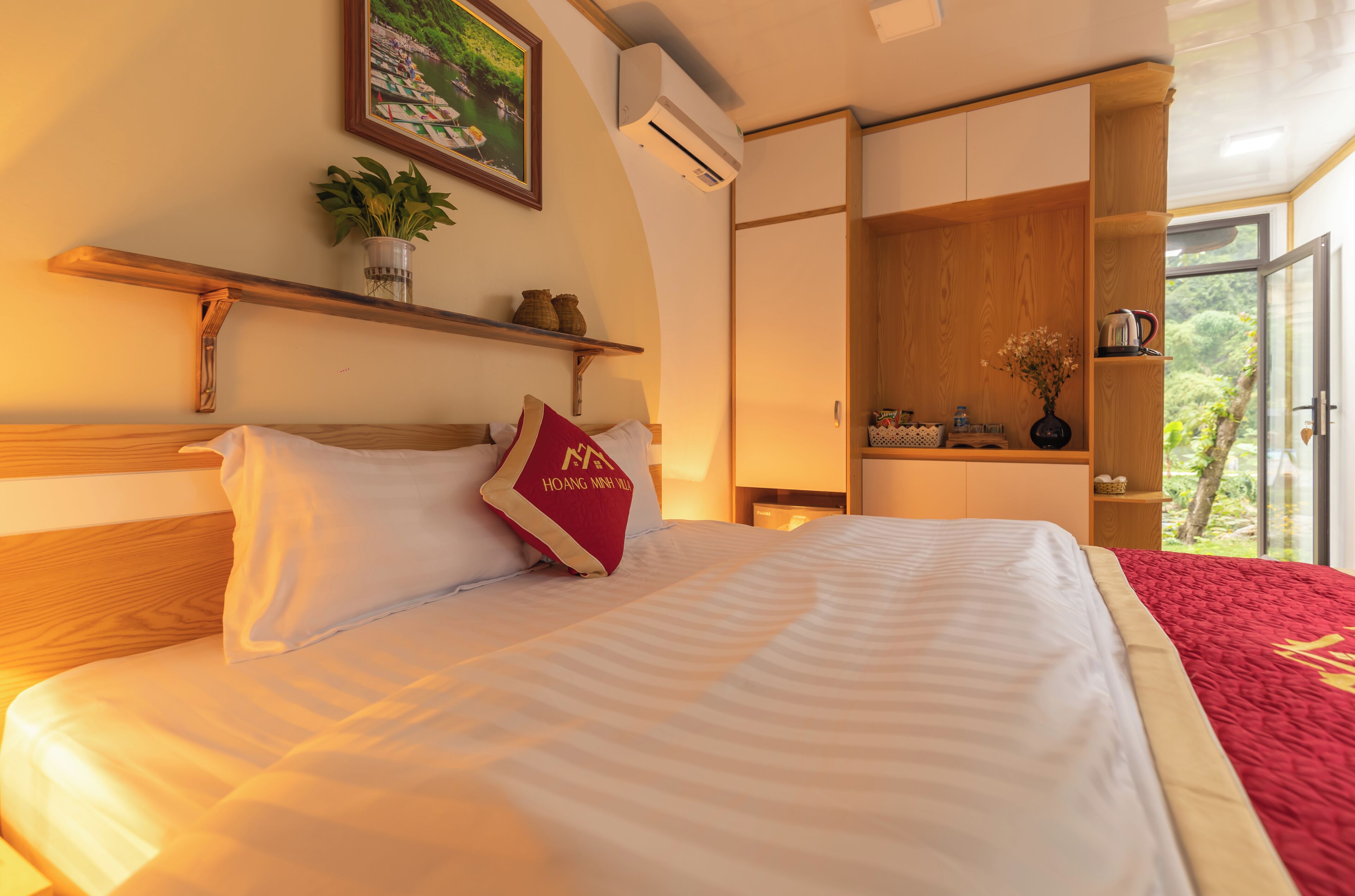 A hotel room with a bed, a red and white pillow, a shelf with a plant, and a window with a view of greenery.