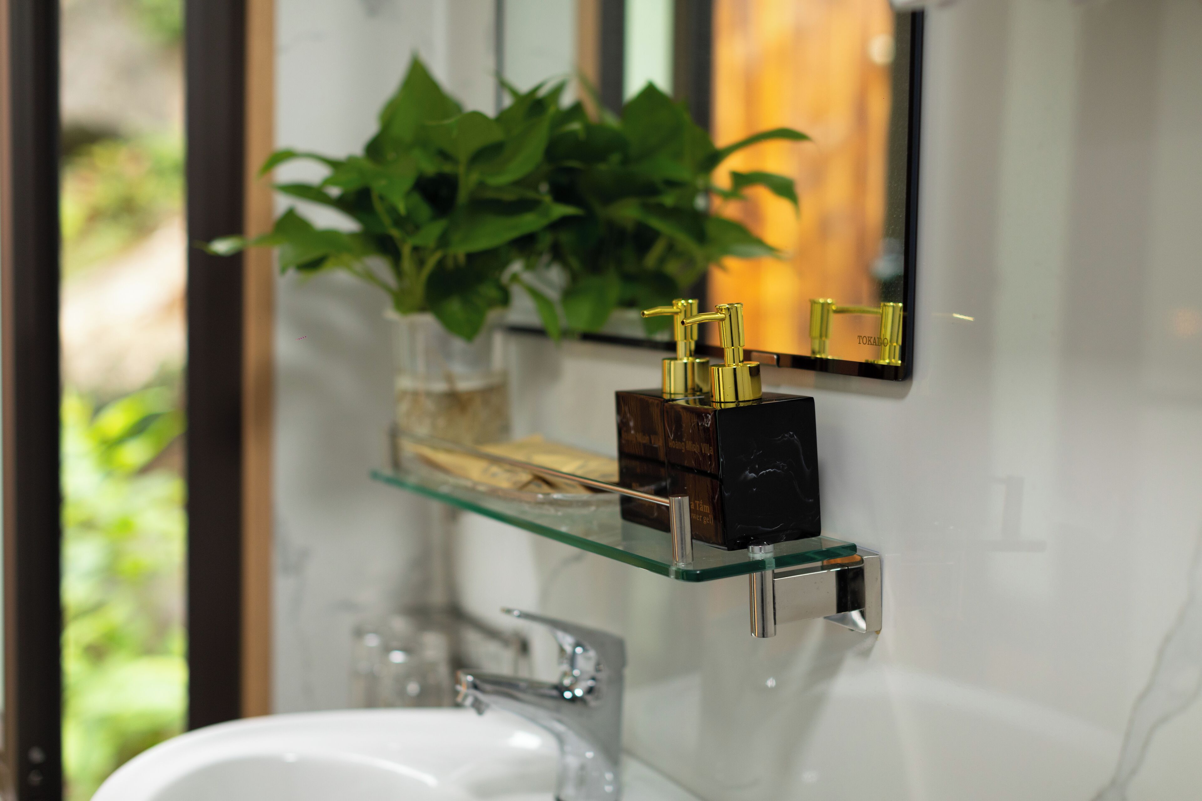 Ein Badezimmer mit Ablagefläche aus Glas, auf der sich eine Pflanze und ein goldfarbener Wasserhahn befinden.