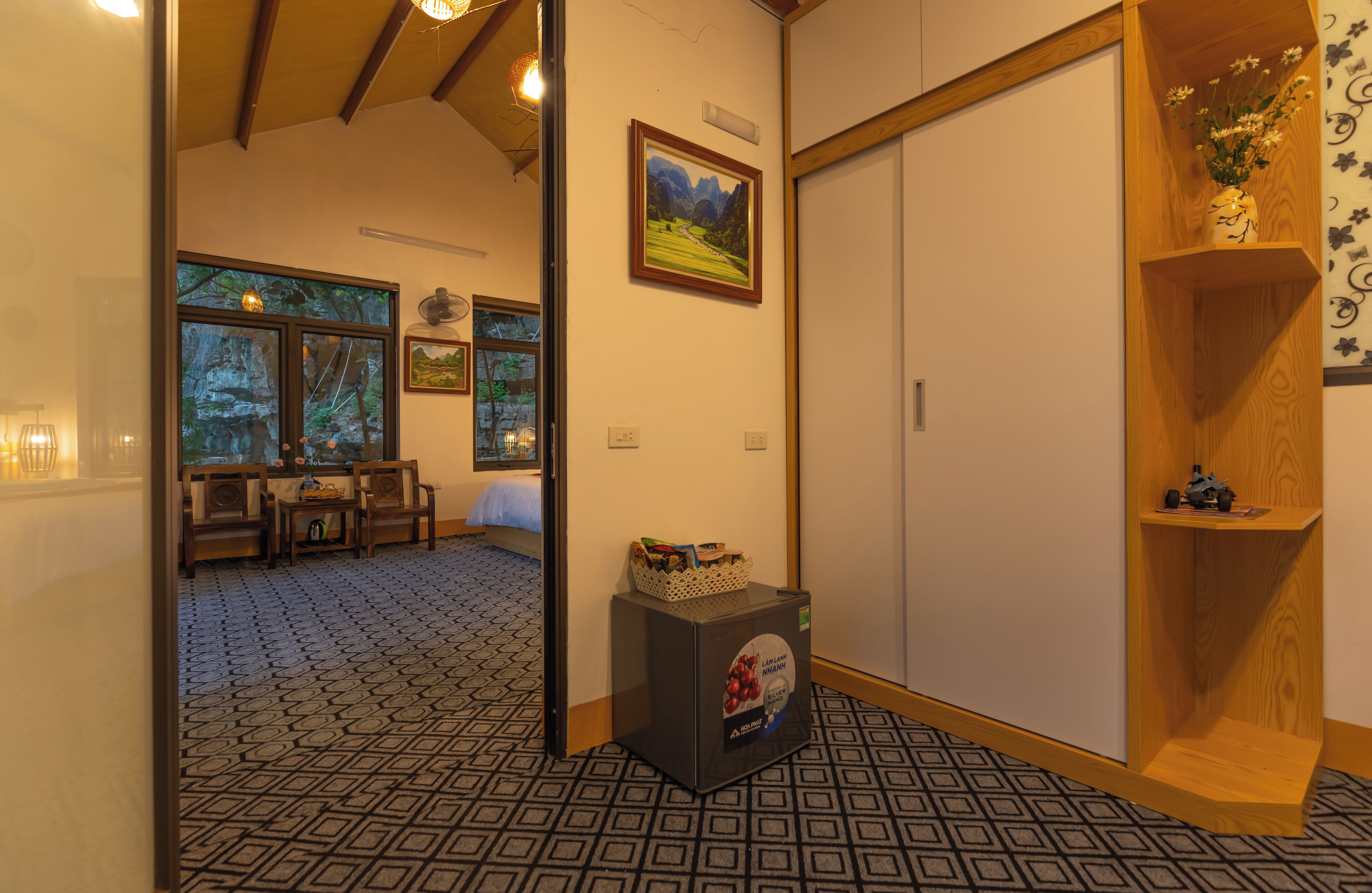 A hotel room with a patterned floor, a wooden cabinet, a framed picture, and a shelf with flowers.