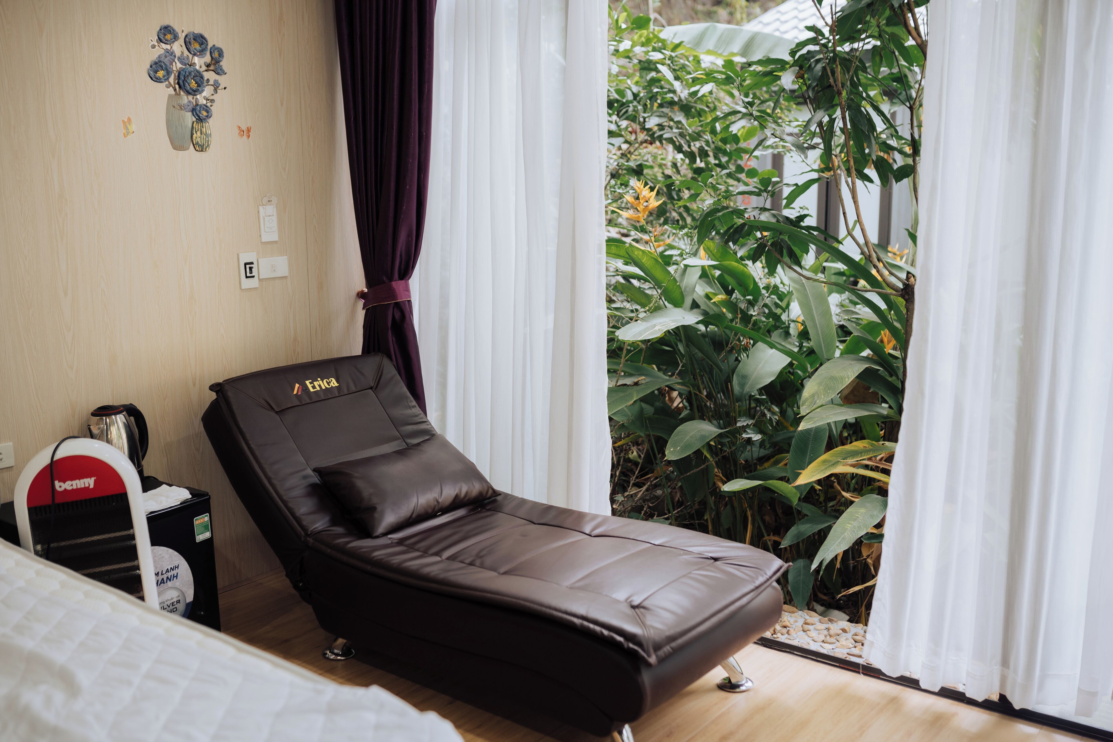A single bed with a white comforter, a brown reclining chair, a small table with a kettle, and a wall-mounted power outlet.
