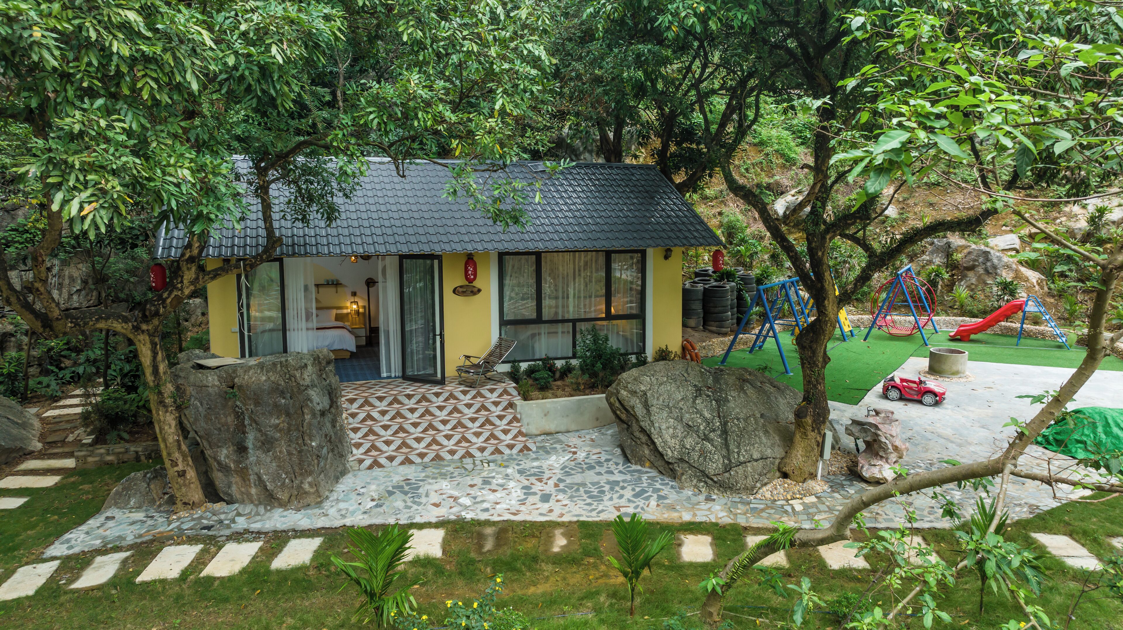 A small house with a tiled patio, a garden with rocks and plants, and a playground with a slide and swings.