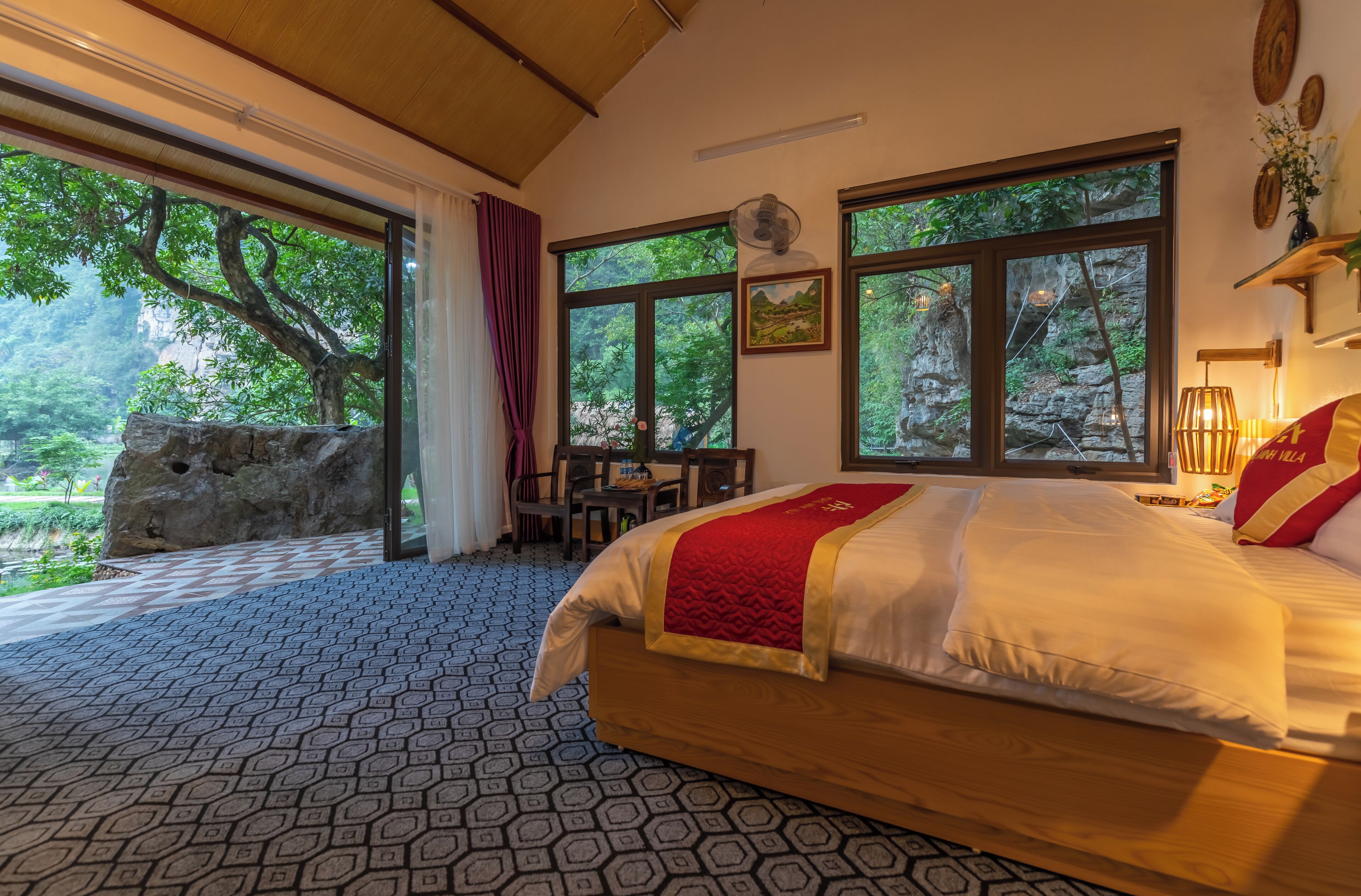 A bedroom with a large bed, a desk, and a view of trees through large windows.