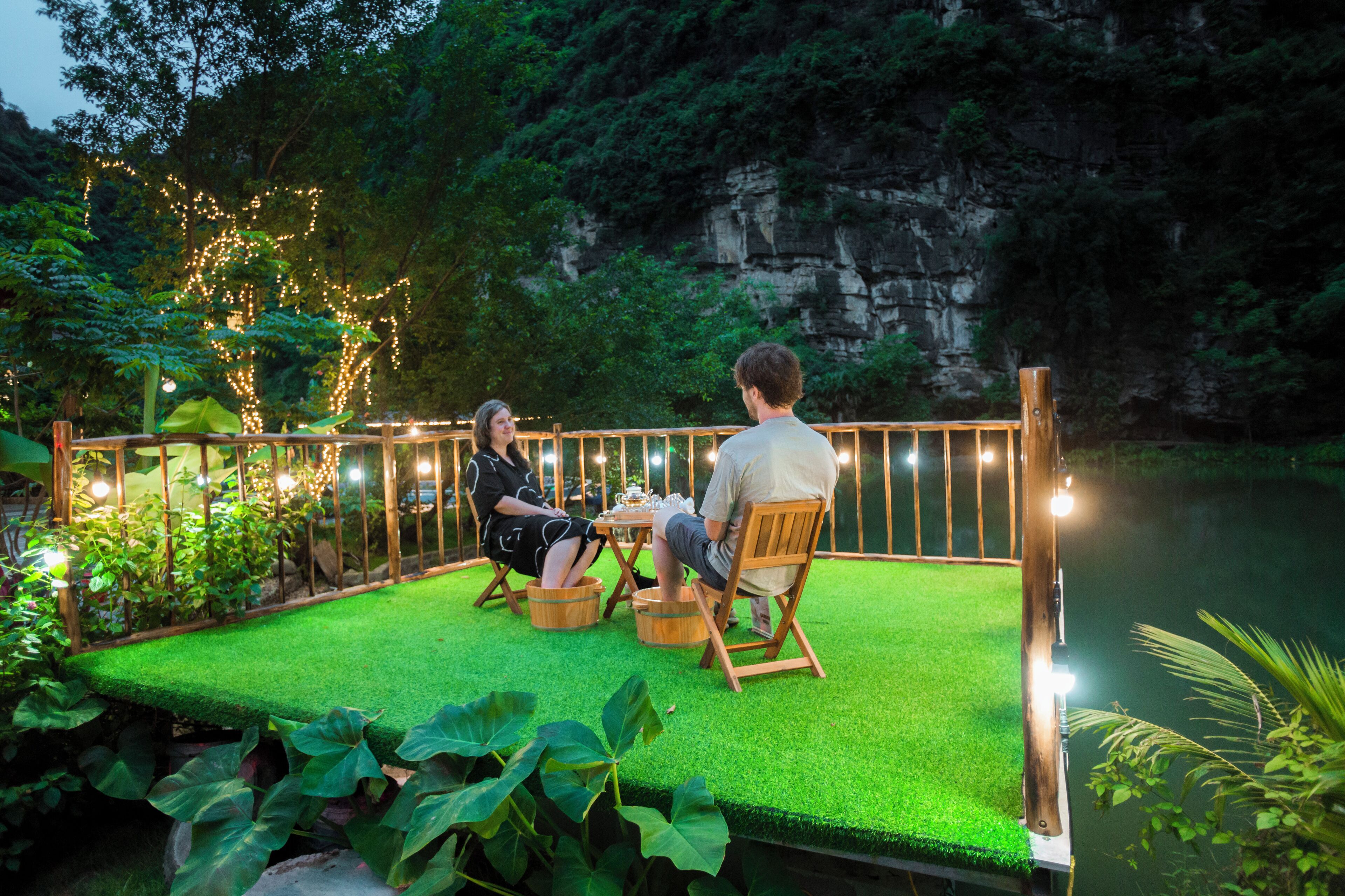 Un couple est assis à une table sur une plateforme recouverte de gazon synthétique vert, avec une rambarde en bois. L’ensemble est entouré de verdure et, en arrière-plan, on aperçoit une falaise rocheuse.