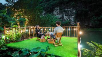 Un couple est assis à une table sur une plateforme recouverte de gazon synthétique vert, avec une rambarde en bois. L’ensemble est entouré de verdure et, en arrière-plan, on aperçoit une falaise rocheuse.