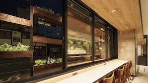 Un interior moderno con una larga barra, sillas de madera y grandes ventanales con vistas a un edificio con plantas.