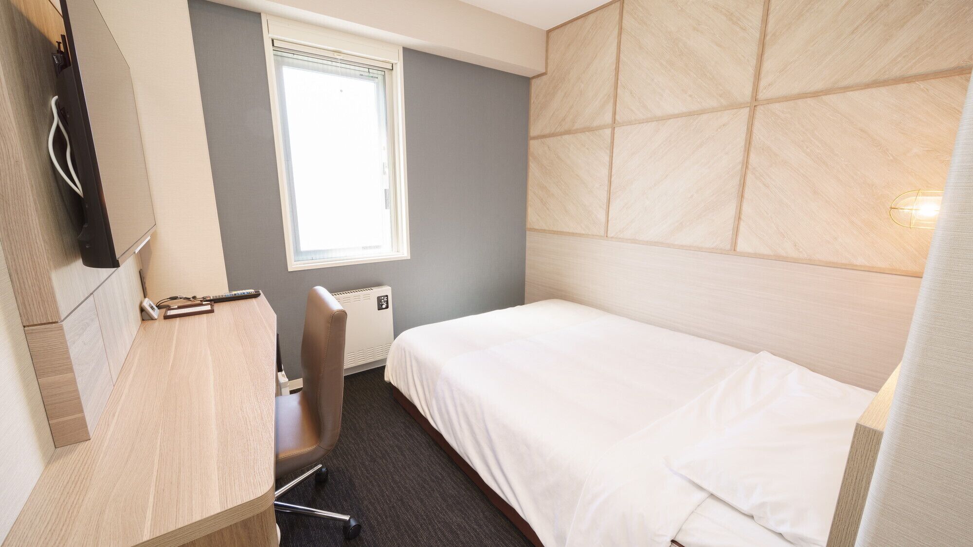 A hotel room with a bed, a desk with a chair, a window, and a wall-mounted TV.