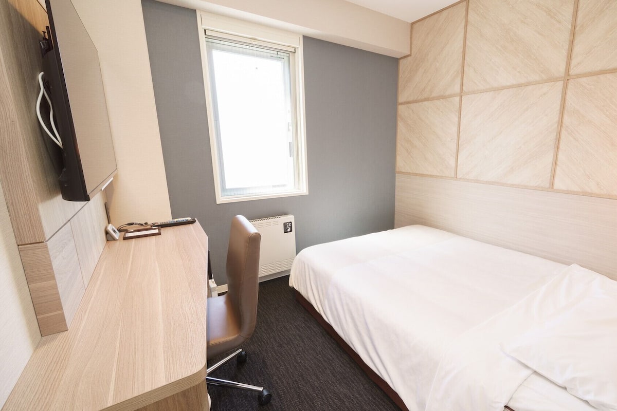 Una habitación de hotel con una cama, un escritorio con una silla, una ventana y un televisor montado en la pared.