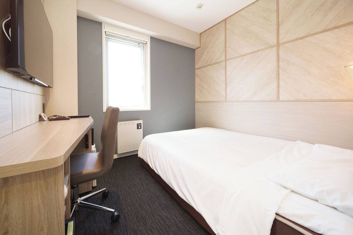 Habitación de hotel con cama, escritorio, silla, ventana y calentador fijado a la pared.