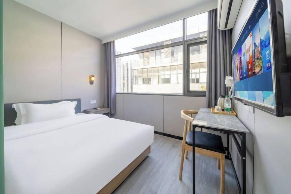 Ein Hotelzimmer mit einem großen Bett, einem Schreibtisch mit Stuhl, einem Fenster mit Blick auf Gebäude und einem an der Wand befestigten Fernseher.