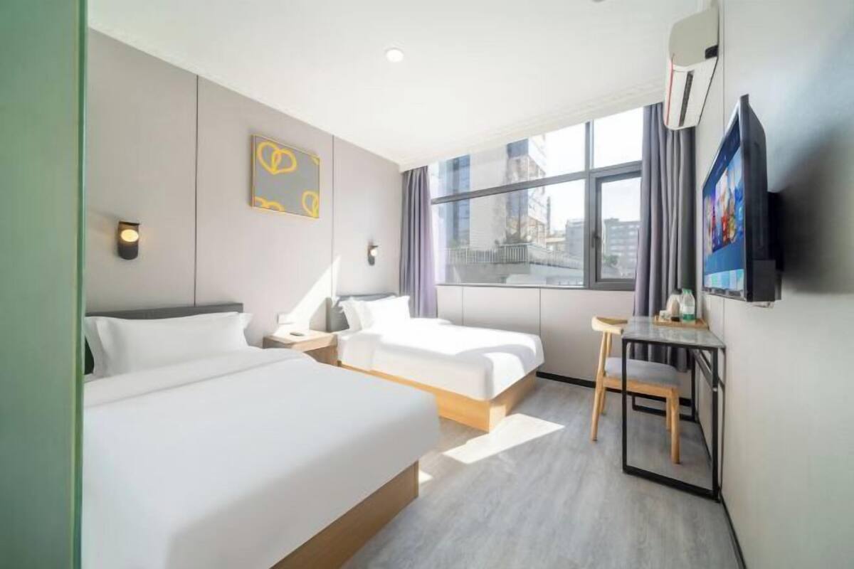 Ein Hotelzimmer mit zwei Betten, einem Schreibtisch, einem Sessel, einem Fernseher und einem Fenster mit Blick auf die Stadt.