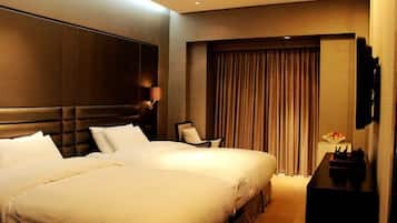 A hotel room with two beds, a large wall-mounted TV, a desk with a lamp, and a large window with curtains.