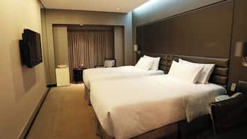 A hotel room with two beds, a TV mounted on the wall, a bedside table with a lamp, and a view of the outside through a window with curtains.
