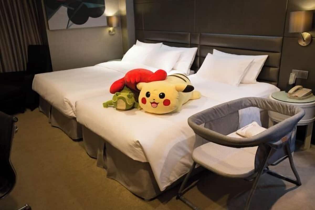 Egy szállodai szoba, amelyben található egy ágy, párnák, egy szék, egy Pikachu mintás párna, egy zöld plüssjáték és egy piros plüssjáték.