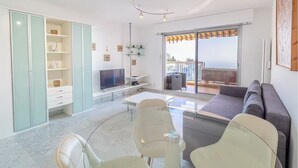 A modern living room with a sofa, TV, and dining area. There is a sliding glass door leading to an outdoor balcony.