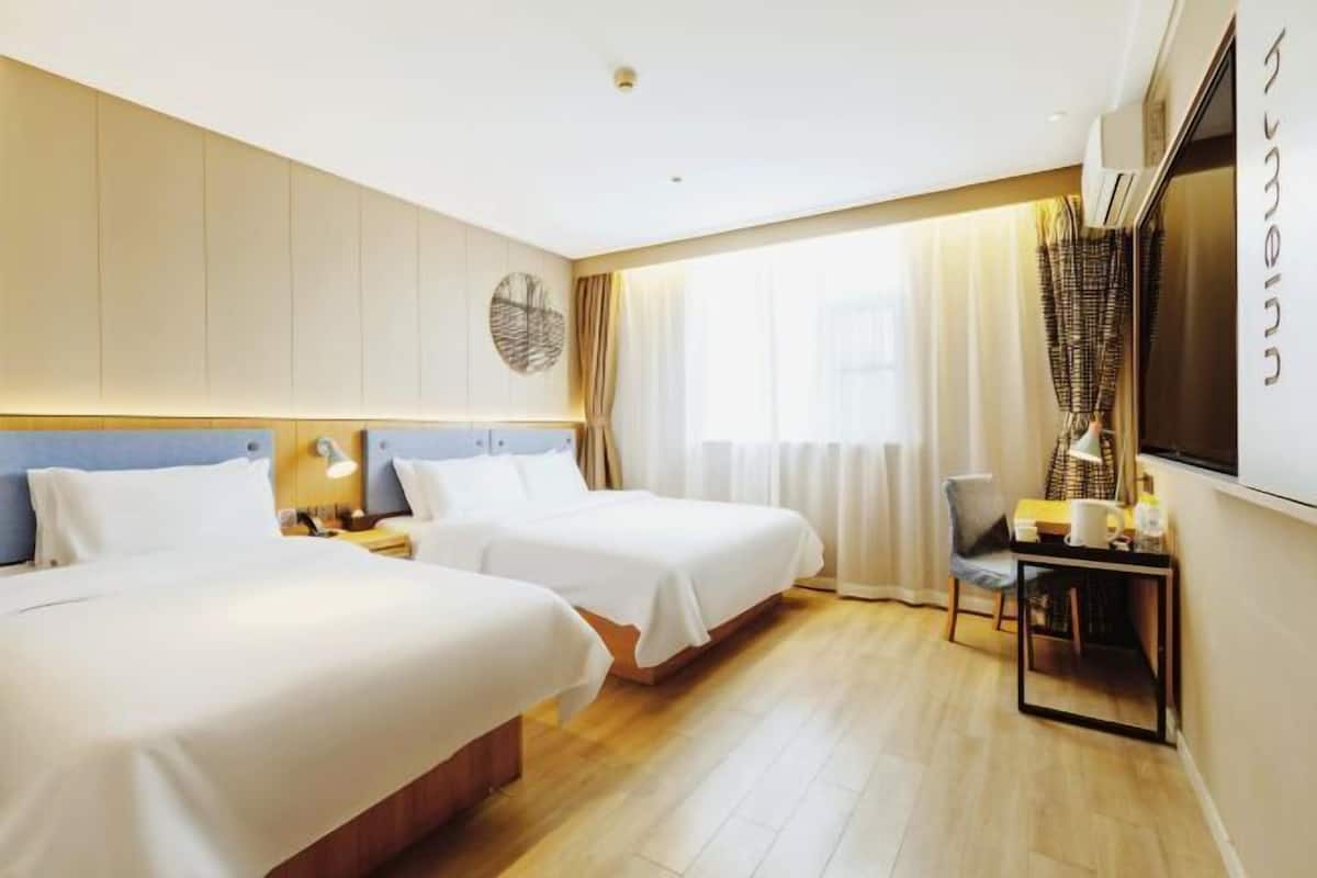 A hotel room with two beds, a desk with a chair, a TV, and a large window with curtains.