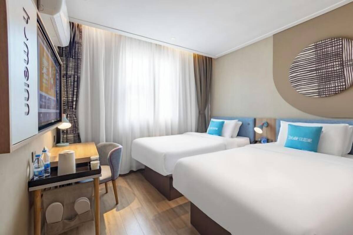 A hotel room with two beds, a desk with a chair, a small table, and a wall-mounted TV.