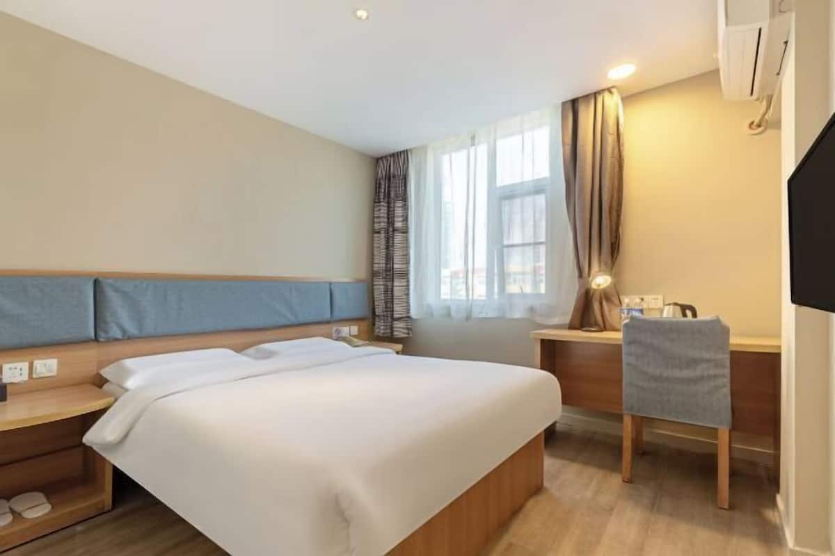 A hotel room with a bed, a desk, a chair, a window with curtains, and a television.