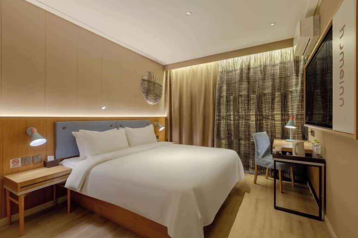 A modern hotel room with a large bed, bedside tables, a desk with a chair, and a large window with curtains.