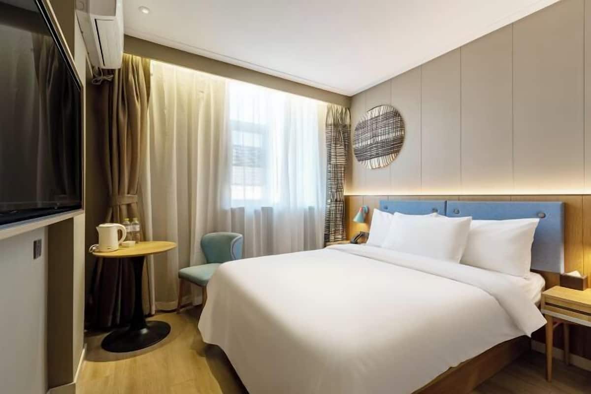 A hotel room with a bed, a small table with a chair, a TV, and a wall-mounted lamp.