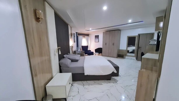 A modern hotel room with a large bed, a bedside table, a wardrobe, and a seating area with a chair and a small table. - Koraf Hotels and Apartments (Abuja)
