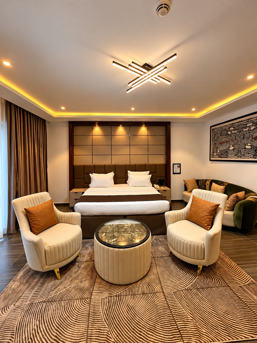 A modern hotel room with a large bed, two armchairs, a coffee table, and a unique ceiling light fixture.