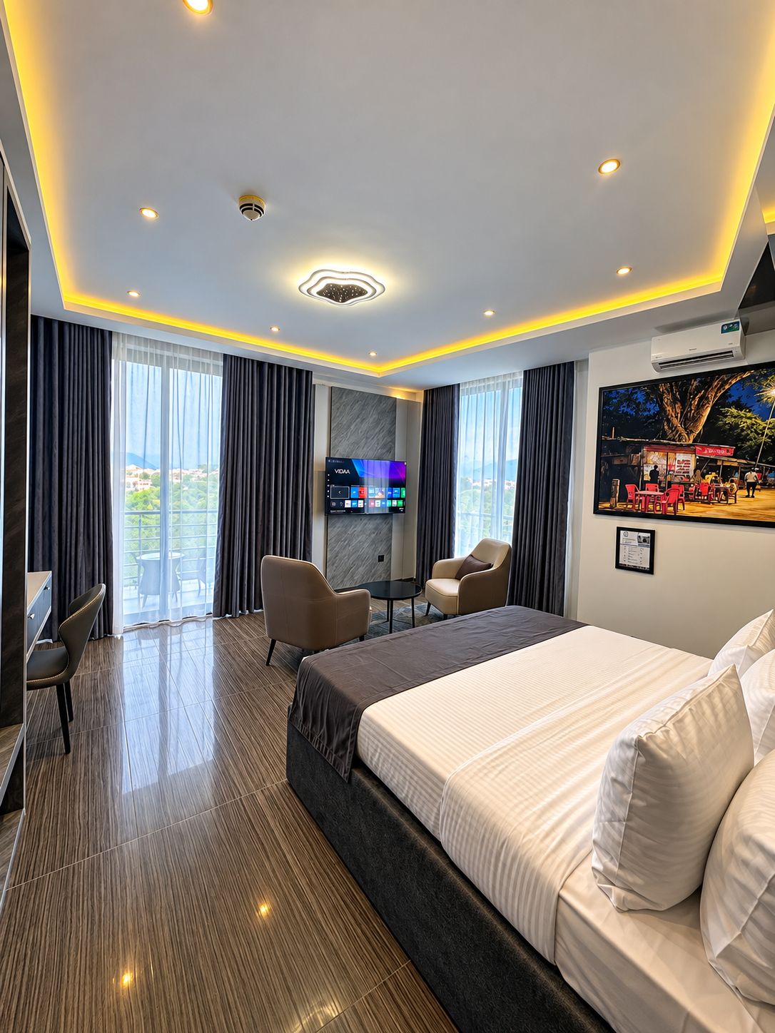 Habitación de hotel con una cama grande, un escritorio, dos sillas, un televisor y un balcón con cortinas.