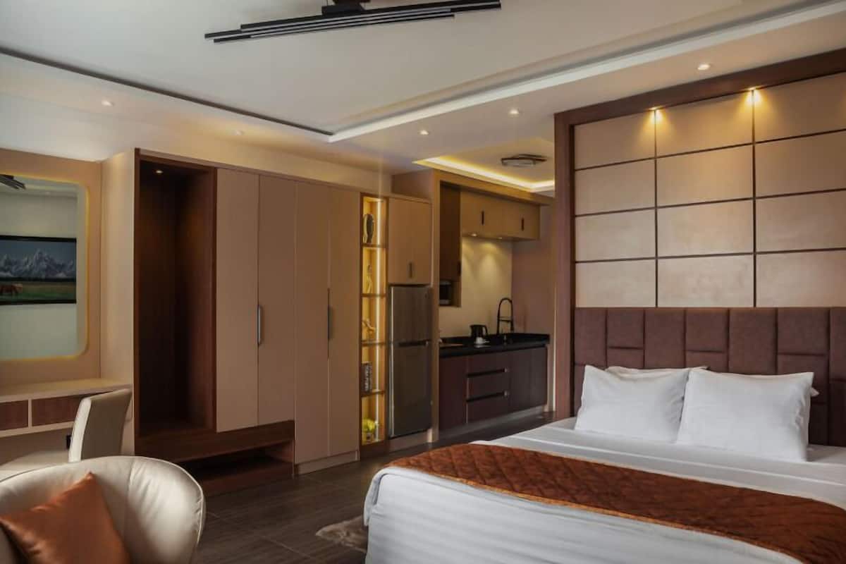 A modern hotel room with a bed, a desk, a chair, a kitchenette, and a large window.