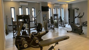 Egy jól felszerelt edzőterem, melyben különféle edzőgépek, egy pad és nagy ablakok találhatók, melyek természetes fényt engednek be.