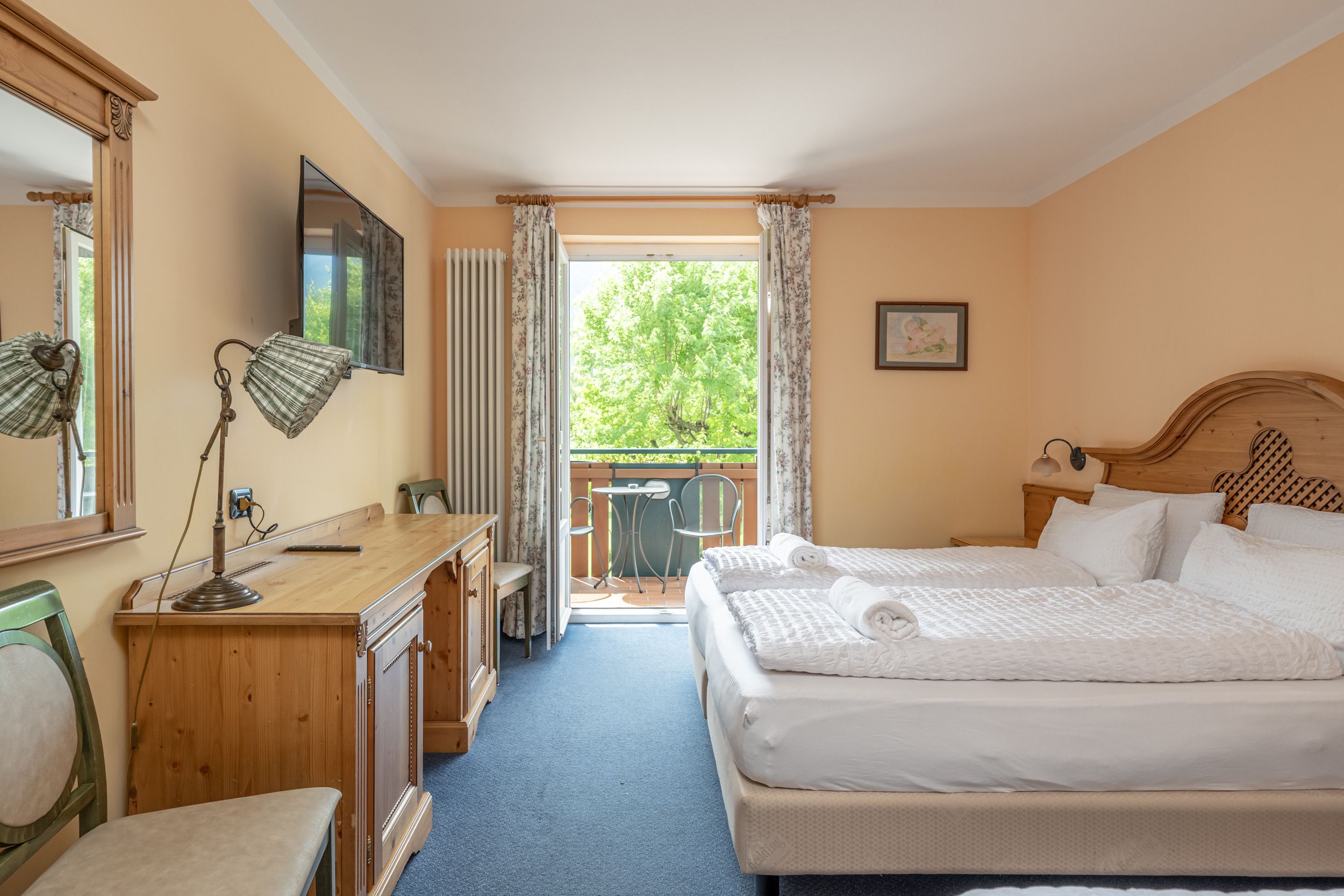Une chambre d’hôtel comprenant un lit, un bureau, une chaise, une commode, un téléviseur et un balcon avec vue sur les arbres.