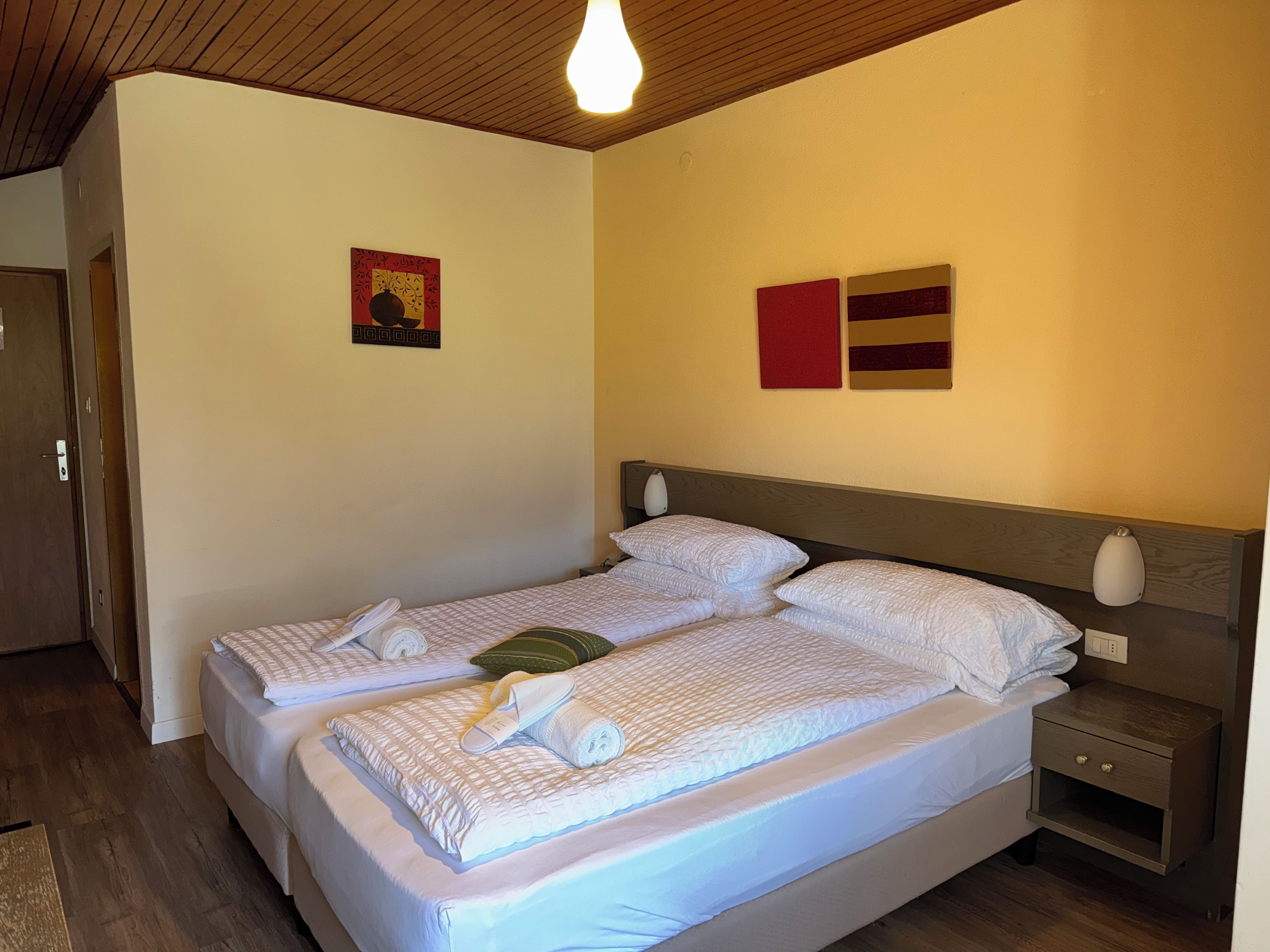 A double bed with two pillows and a folded towel, a bedside table with a lamp, and a wall with two framed artworks.