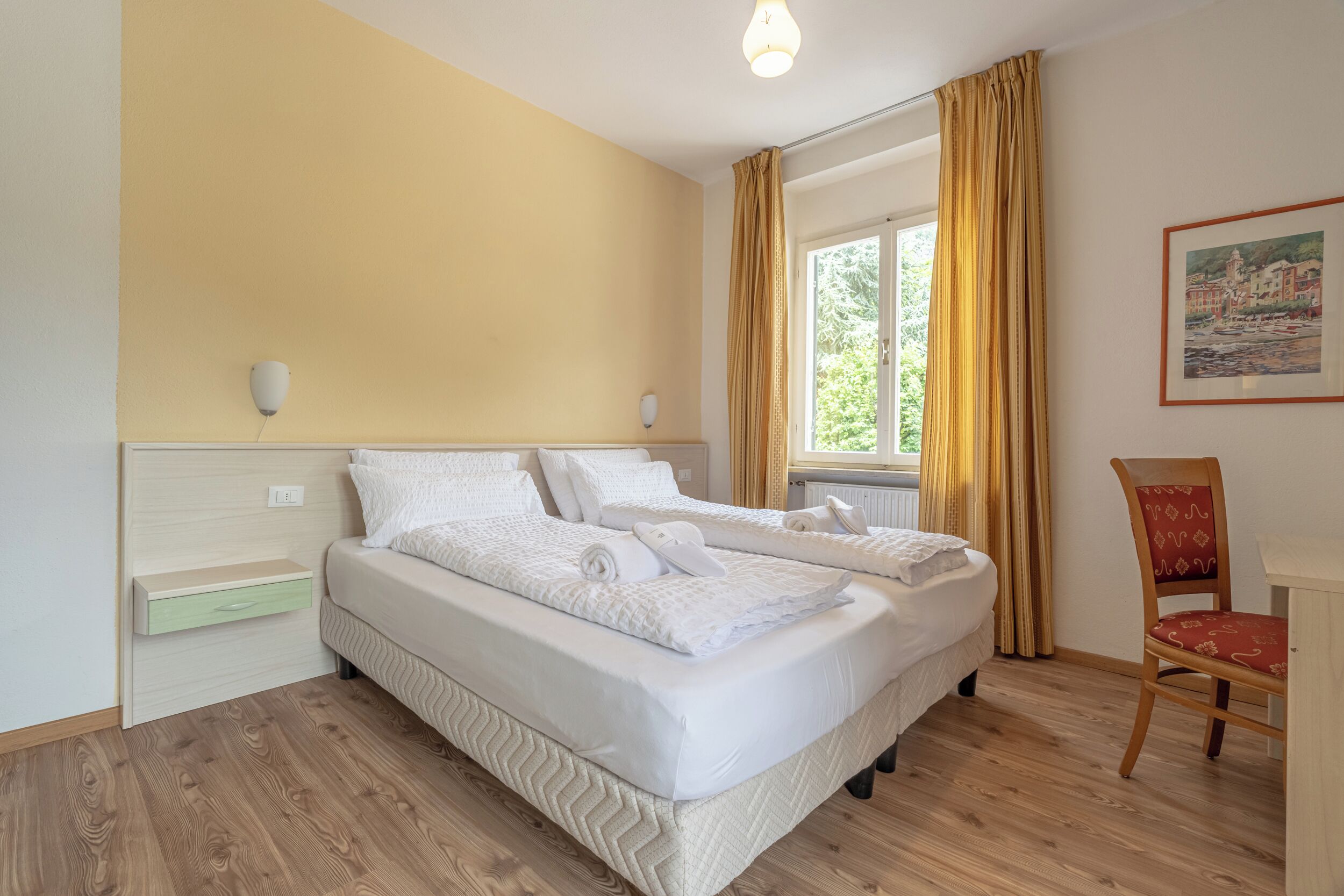 Une chambre d’hôtel avec un lit, une chaise, une fenêtre avec des rideaux, un luminaire fixé au mur et un tableau.