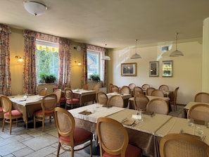 A dining area with multiple tables and chairs, floral curtains, and wall-mounted lights. - Garden Hotel Bellariva (Molveno)