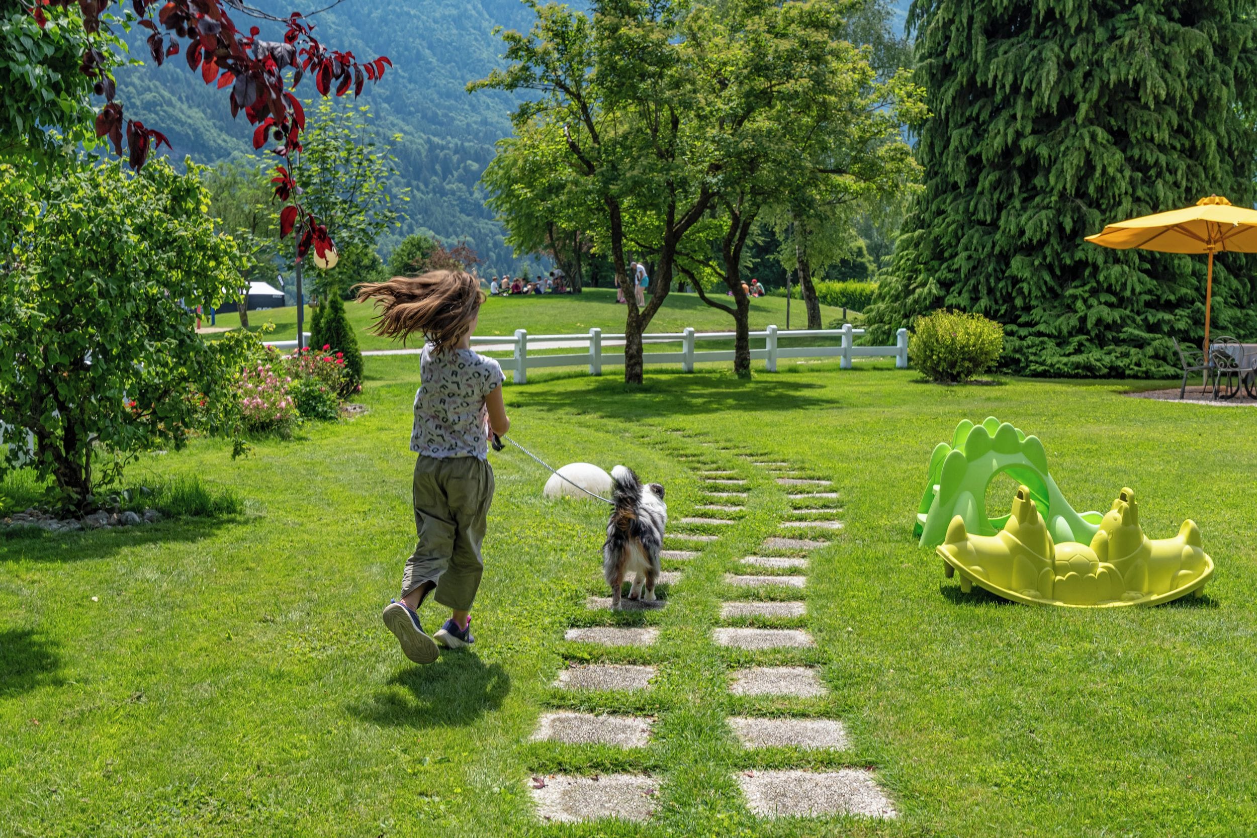 Eine Person spaziert mit einem Hund auf einem gepflasterten Weg in einem Park mit üppiger Vegetation und einem Berg im Hintergrund.