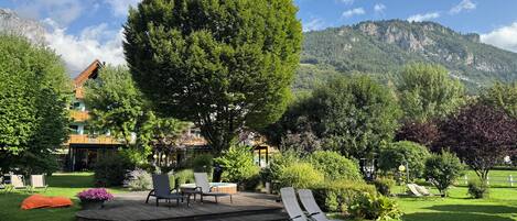 A lush garden with a large tree, outdoor seating, and a mountain backdrop.