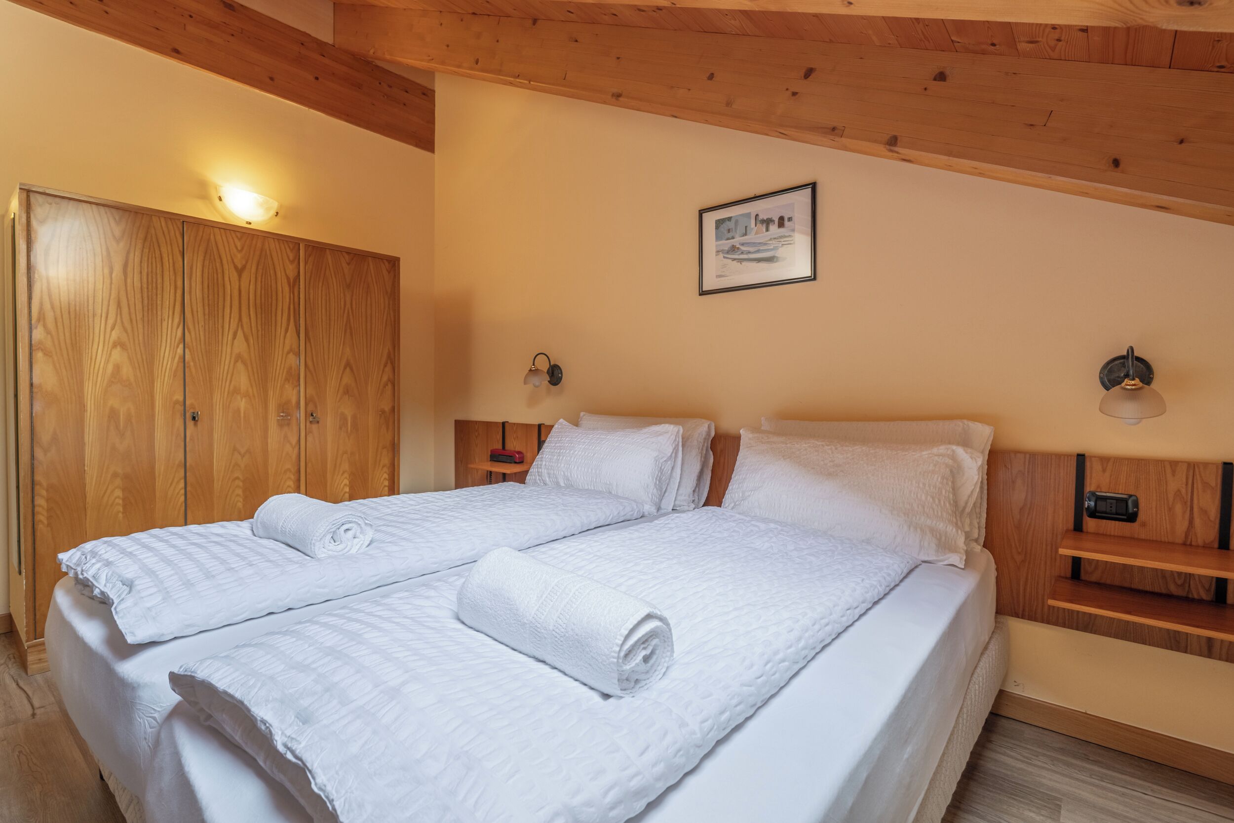 A double bed with white bedding and rolled towels, wooden wardrobe, wall-mounted light fixtures, and a framed picture on the wall.