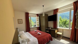 A hotel room with a bed, desk, and large windows offering a view of greenery.