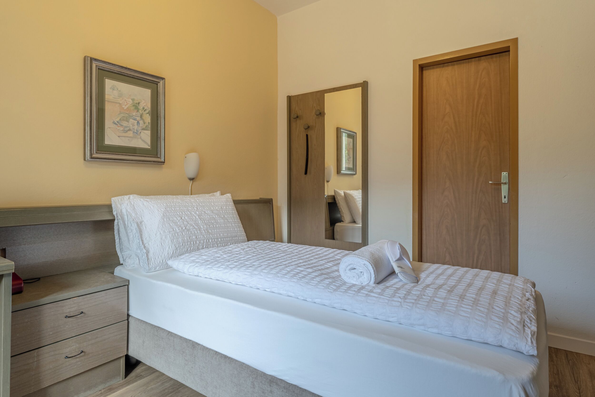 A neatly made bed with a white duvet and pillows, a bedside table with a lamp, a wooden headboard, a framed picture on the wall, and a wooden door.