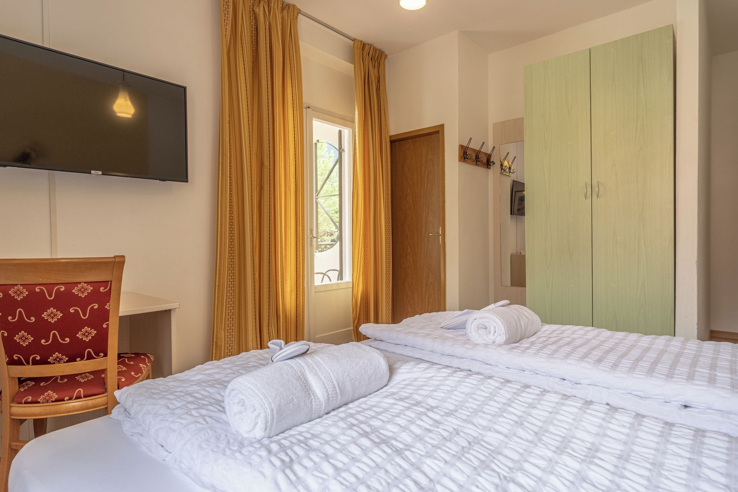 A hotel room with two beds, a TV mounted on the wall, a wooden chair, and a wardrobe.