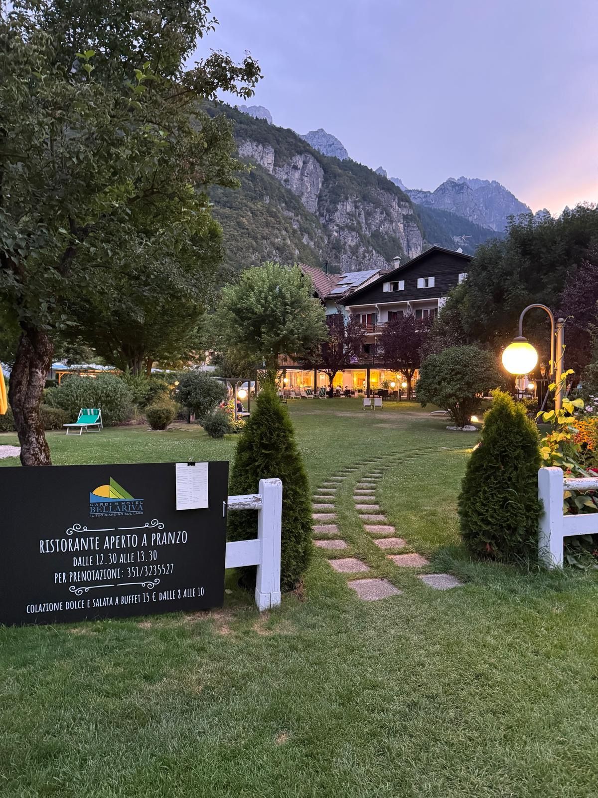 Ein Gartenhotel mit einem Schild, das ein Restaurant im Freien ankündigt, einem Berg im Hintergrund und einem gepflegten Rasen.