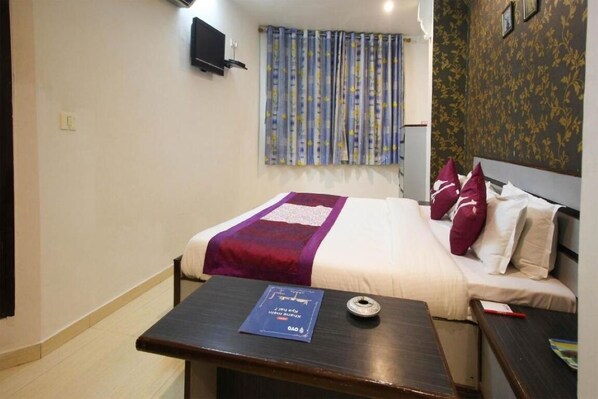 A hotel room with a bed, a bedside table with a book, a wall-mounted TV, and a window with curtains. - Hotel Liberty (Ahmedabad)