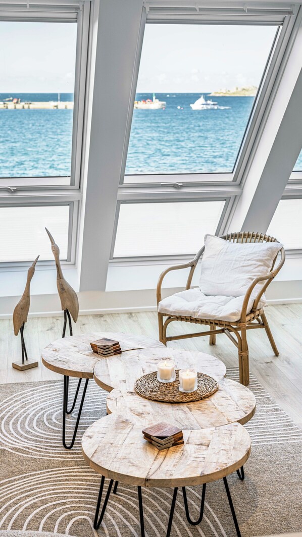 A room with a large window overlooking the sea, featuring a wicker chair, a round wooden table with candles, and decorative bird sculptures. - Villa sea view of Roscoff - Sauna (Roscoff)